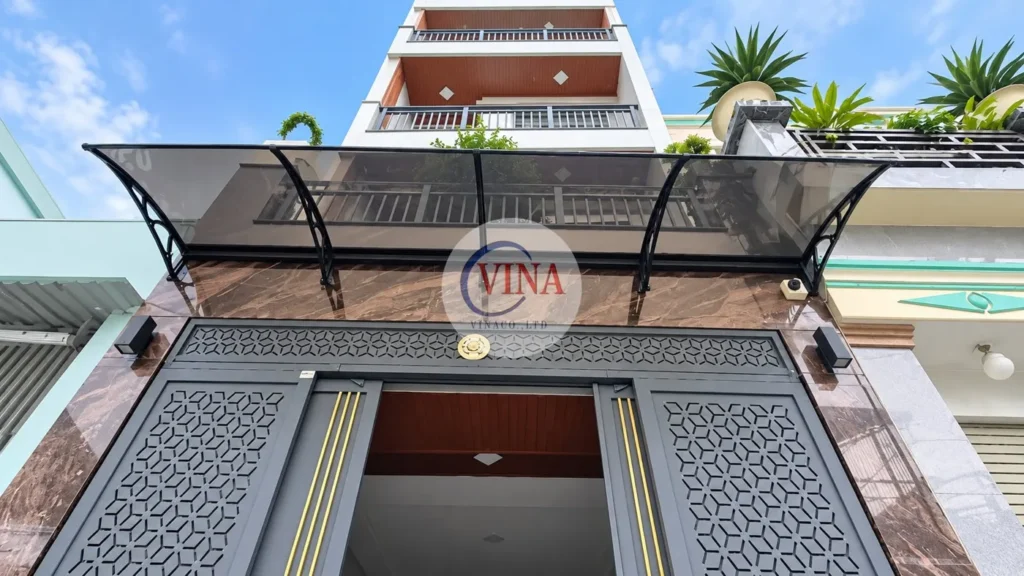 Chính diện mặt tiền: mái che canopy tấm poly nâu khói với 4 tay đỡ đen, gắn trên mảng tường ốp đá phía trên cửa cổng.