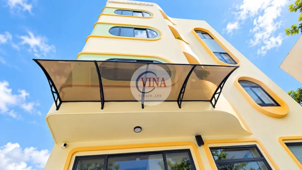 Chính diện: mái che canopy tấm poly nâu khói với 4 tay đỡ đen, gắn trên tường tầng lầu, ngay phía trên phần sảnh bo cong của tầng trệt.