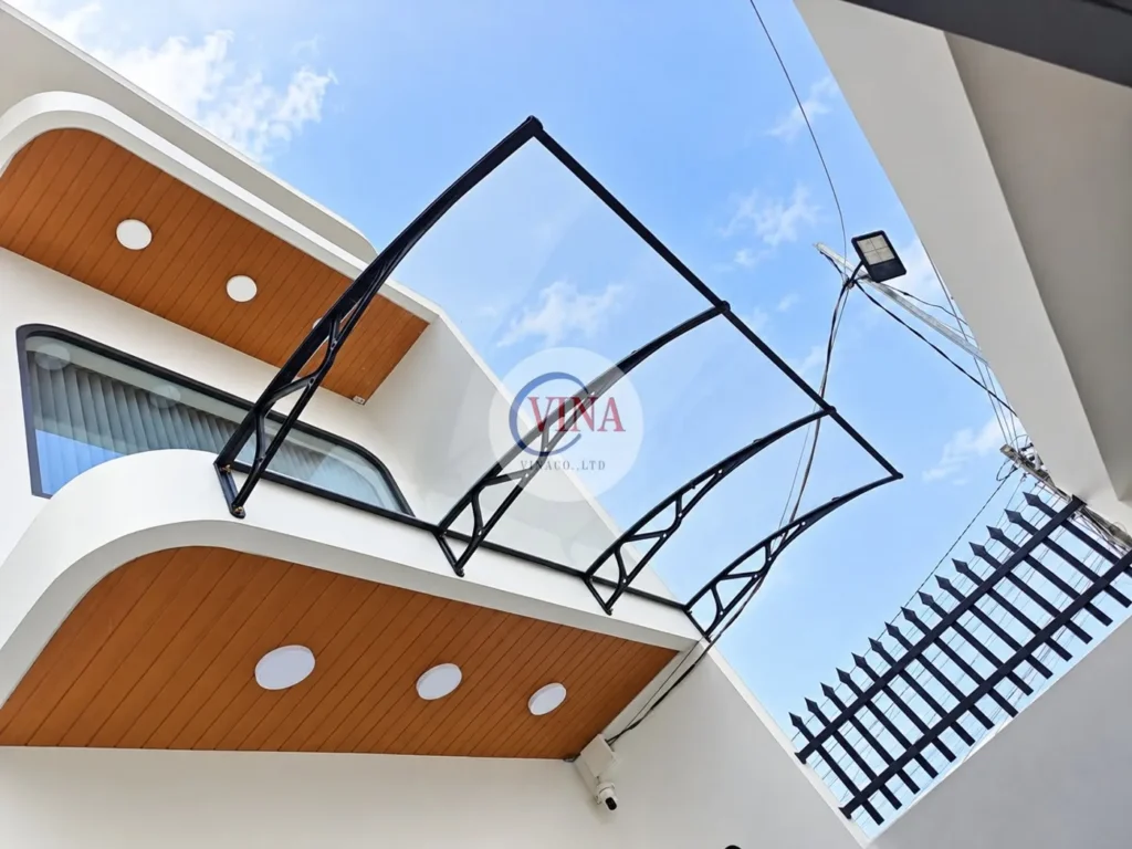 Mái che canopy tấm poly trong suốt, 4 tay đỡ đen, gắn trên tường tầng lầu giữa hai khối bo cong có trần ốp vân gỗ.