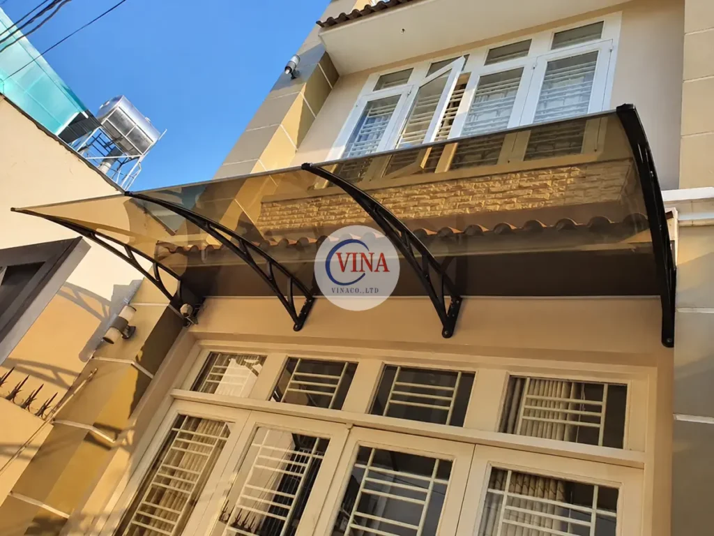 Mái canopy màu nâu trà che cửa ra vào, tay đỡ đen nổi trên tường vàng nhạt.