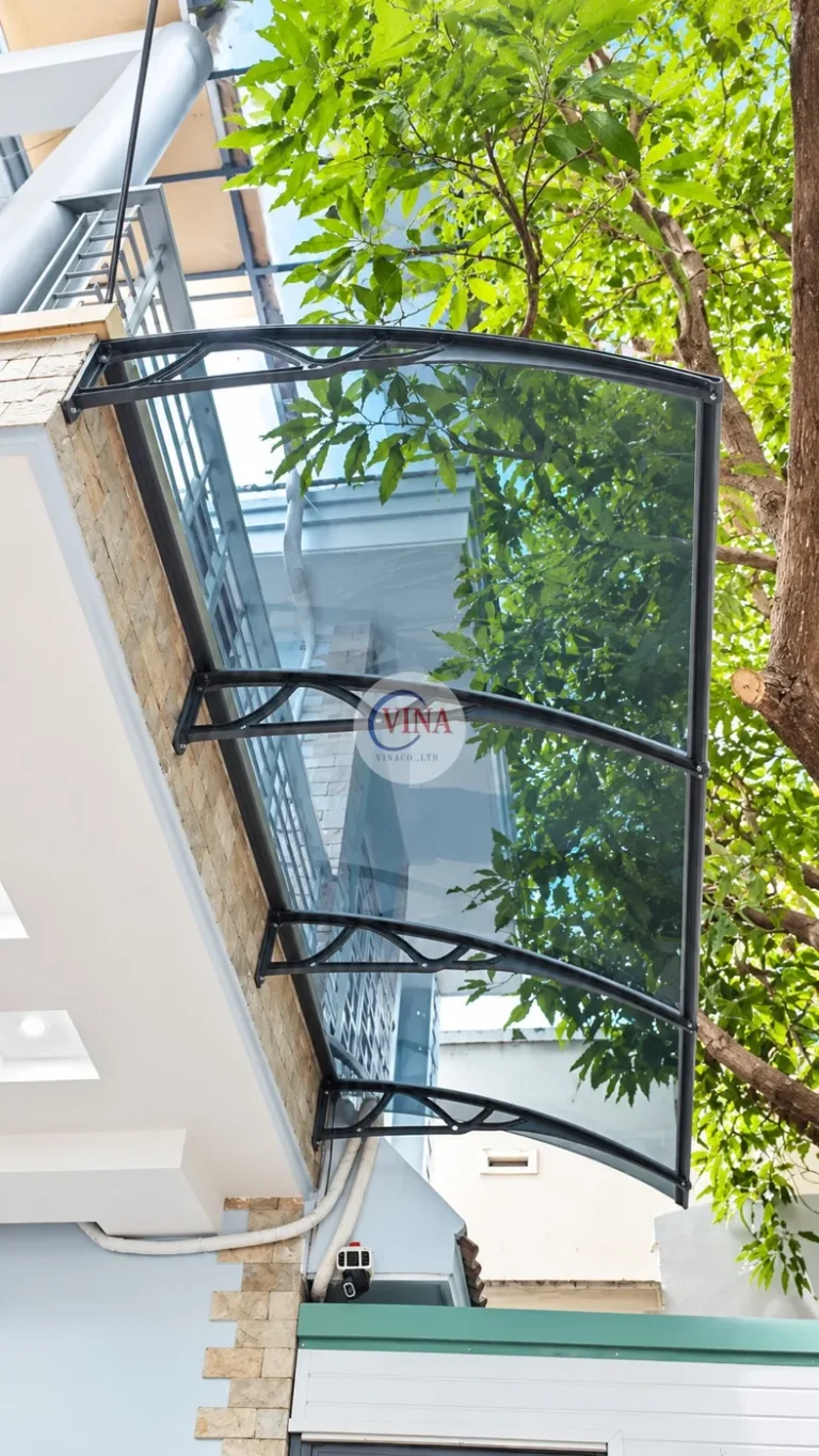 Nhìn từ dưới lên: tấm polycarbonate xám khói uốn cong theo tay đỡ, vẫn thấy rõ tán cây và bầu trời phía trên.