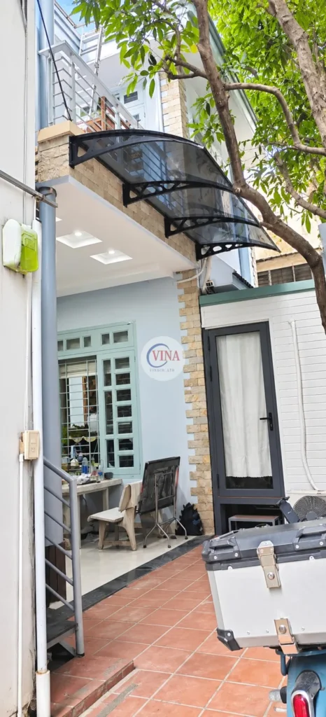 Toàn cảnh: mái che canopy tấm poly xám khói lắp ở mép sàn ban công tầng lầu, vươn ra che khoảng sân lát gạch phía trước nhà.